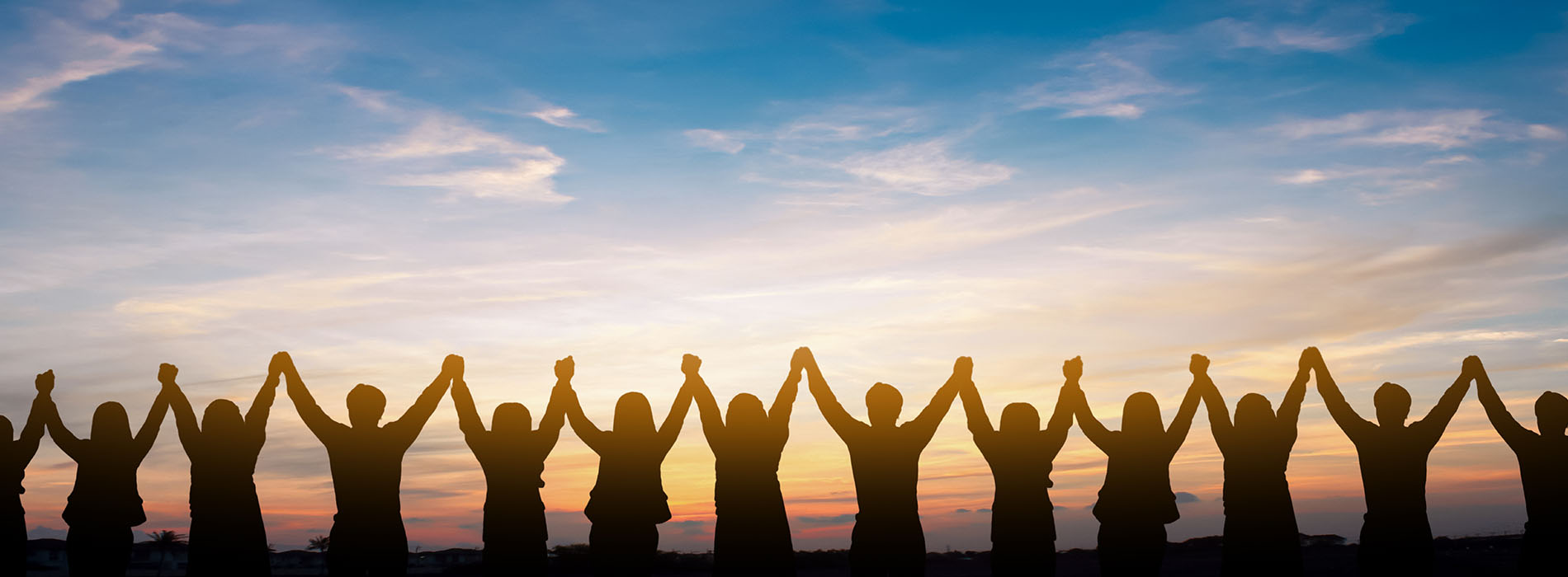 The image shows a group of people standing together with their hands raised, creating a silhouette against a sunset sky, all facing the same direction.