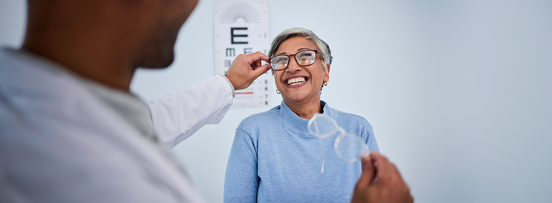 The image shows a medical professional assisting an elderly person during an eye examination, with the patient seated and the professional standing to the side, holding up a chart.