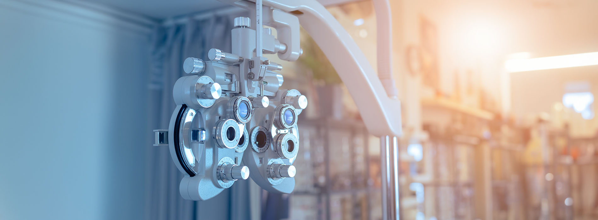 The image shows a close-up view of an optical machine with various lenses and components, placed against a blurred background that appears to be an indoor setting with shelves and items on display.