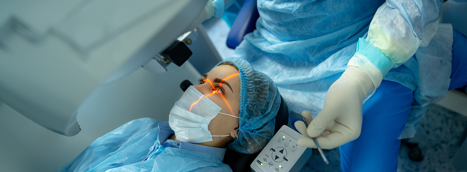 The image shows a medical setting with a person wearing blue scrubs, a surgical mask, and gloves, performing a procedure on a patient lying on an operating table. A surgeon s hand is visible holding a scalpel, and there appears to be a monitor displaying medical images in the background.