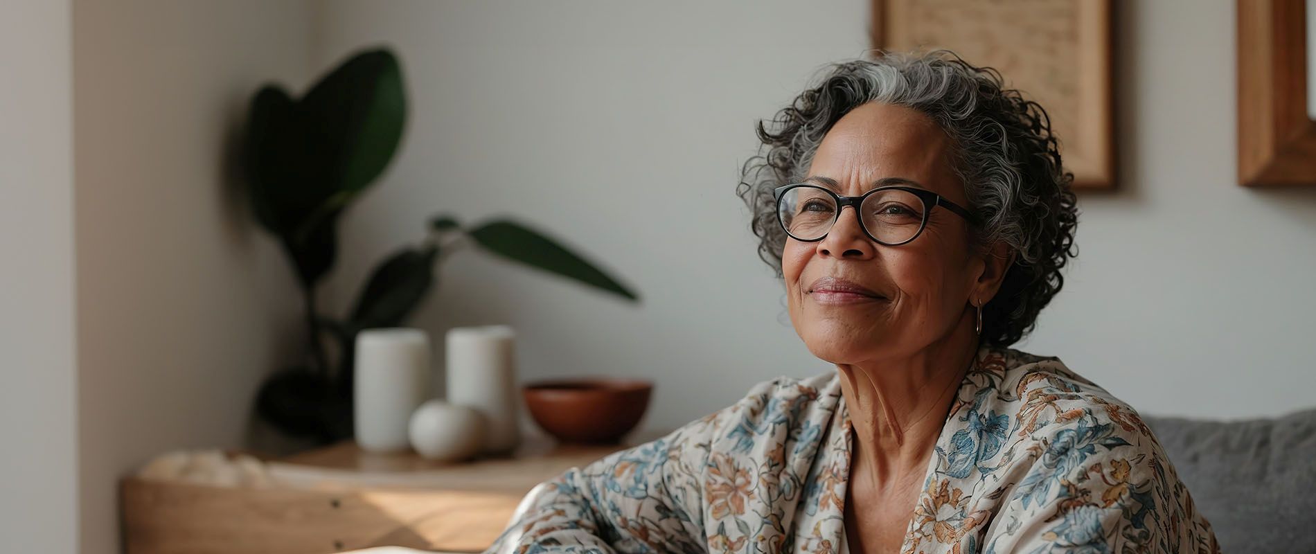 An elderly woman with glasses sits in front of a houseplant, looking out a window with her hands clasped together.