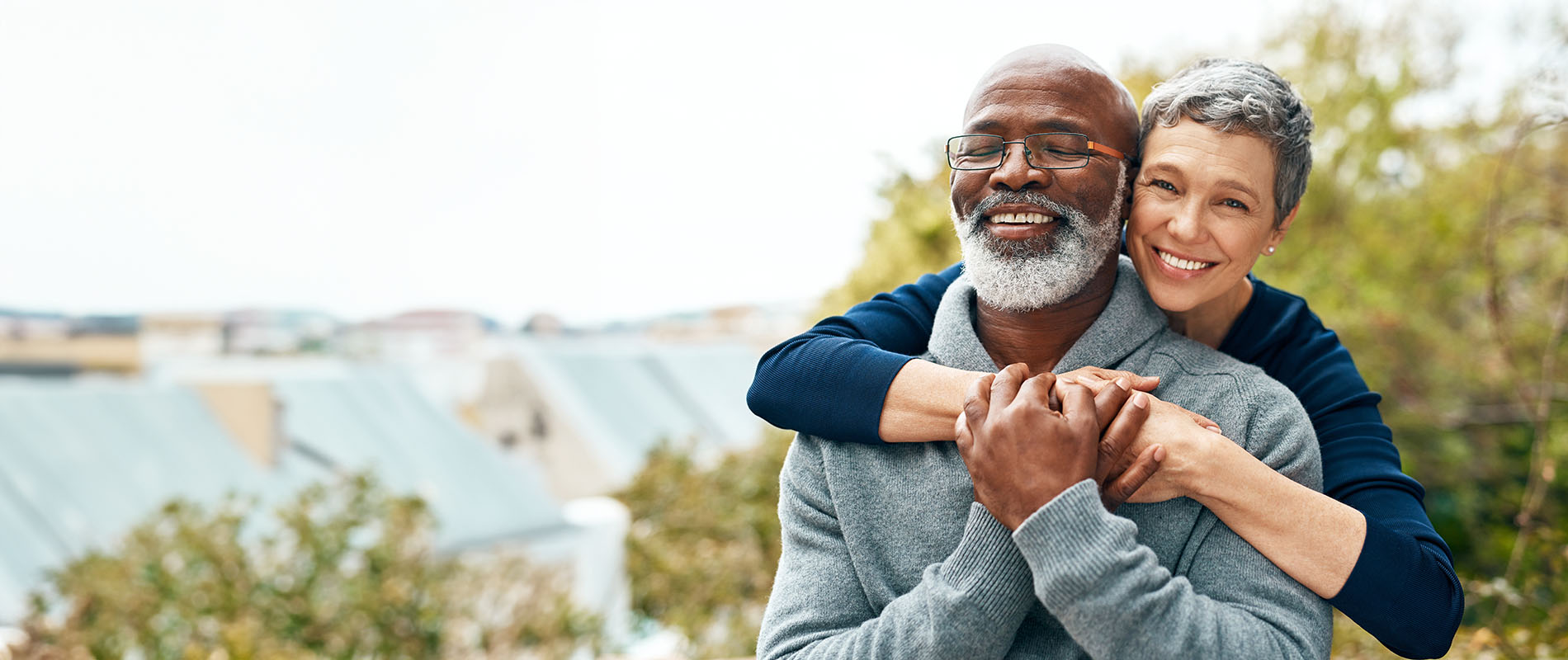 The image shows two people, likely an older couple, embracing each other with smiles on their faces, set against a blurred background featuring greenery and a building.
